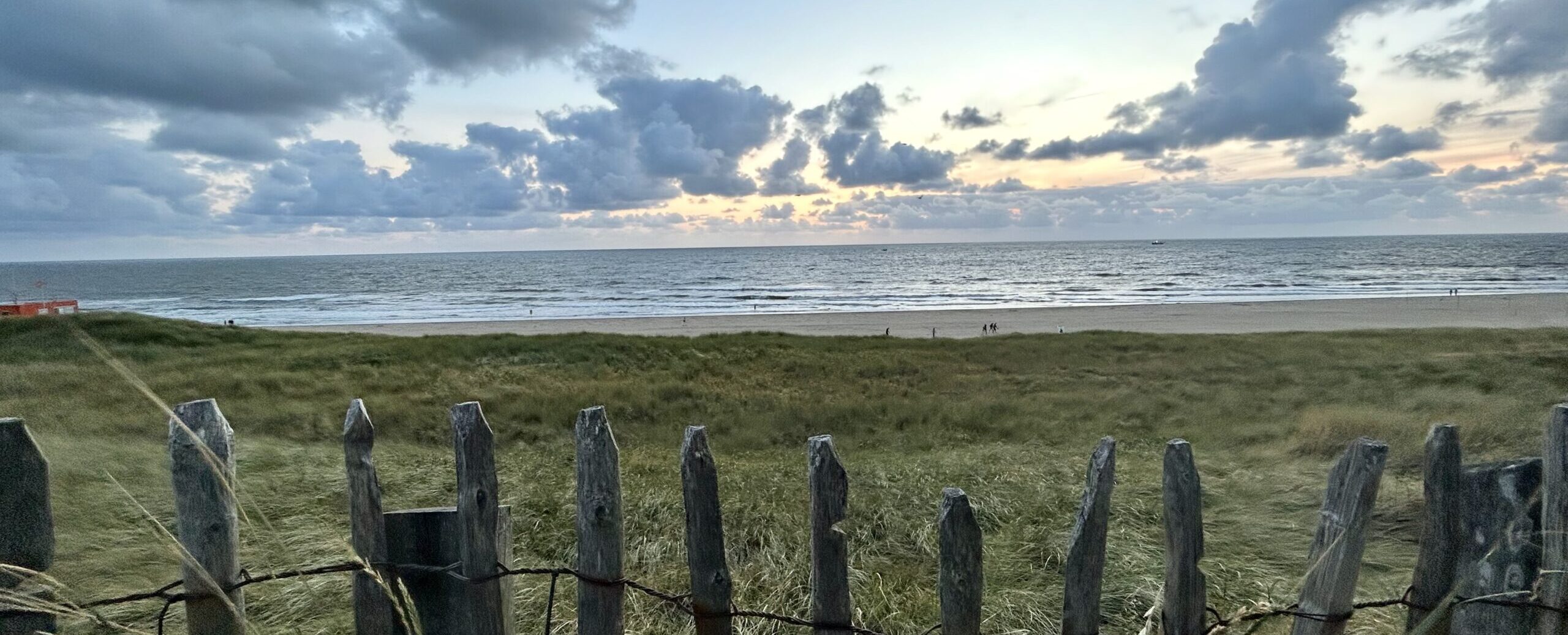 Een houten hek in de duinen, met strand en zee erachter.